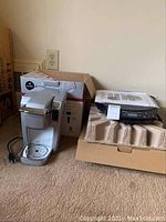 Keurig coffee maker and NuWave induction cooktop side by side on carpet with boxes.