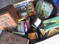 Overview of large bin filled with assorted antique branded tins and boxes showing visible wear including rust and scratches.