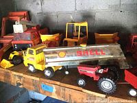 Photo showing a Shell chrome tanker truck, red tractor, yellow dump trucks, and orange construction vehicles all on a wooden shelf.