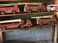 Five vintage used red toy fire trucks displayed on wooden shelves showing ladders and decals with wear and rust.