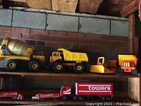 Photo showing seven vintage toy trucks including a Tonka cement mixer, dump truck, orange bulldozer, and red Towers branded trailers, all aged with wear and surface imperfections.