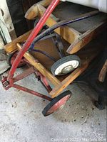 Side view of wooden child wagon with red metal wheels and handle, showing general condition.