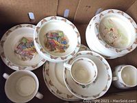 Group of Royal Doulton Bunnykins ceramic dishware in a box showing bowls, plates, cups, and saucers with bunny border motifs and bunny/child scene illustrations.