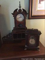 View of both antique clocks on a wooden surface. Taller clock with finial at back left, smaller clock at front right.