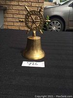 Front view showing brass ship's bell with detailed ship wheel handle in a golden polished finish.