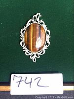 Front view of the large ornate pewter pin with polished oval tiger eye cabochon on green background with lot number card.