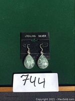 Pair of sterling silver earrings with teardrop-shaped petersite stones displayed on a black card reading 'STERLING SILVER'. Green background beneath. Lot number 744 shown in front.