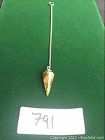 Full view of the tiger's eye pendulum with the metal chain against a green background, showing the length and the pointed shape of the stone pendulum.