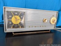 Front left angled view of vintage Admiral 5W3X tube clock radio showing yellow and black clock face with two knobs and Admiral metal lettering on grey horizontal ridged steel casing. Two tuning knobs on right side with large numbered dial.
