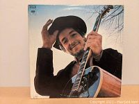 Front cover of Bob Dylan's Nashville Skyline album showing Bob Dylan with an acoustic guitar and wearing a hat, smiling at the camera.