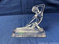 Side angled view of the clear crystal paperweight showing the figure in seated pose with arm behind head against a dark blue fabric background.