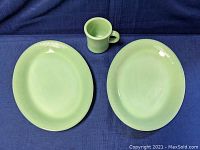 Top down view of two oval Fire King Jadite plates and one coffee mug on blue cloth background. Plates measure approximately 12 x 9 inches.