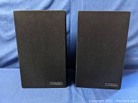 Front view of two Mission Model 70 speakers covered with black fabric grills, showing the Mission logo bottom right.