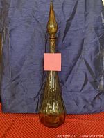 Front view of tall amber Empoli glass decanter with elongated neck and teardrop-shaped stopper against purple and red background.