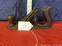 Side view of vintage woodworking hand plane on red patterned surface with measuring tape in background showing length approximately 14 inches .
