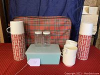 All items in the vintage picnic set displayed on a red tablecloth with a plaid insulated bag in the background.