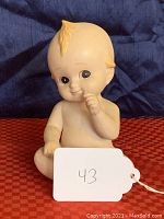 Front view of bisque porcelain Kewpie doll by Lefton sitting with one hand near mouth.