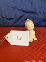 Front view of a seated bisque porcelain Kewpie doll figurine with a finger near its mouth against a red checkered surface and blue background, showing some wear.