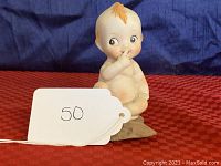 Kewpie doll facing front with finger to mouth, sitting on small stone base with glue visible, placed on red fabric background.
