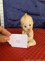 Front view of bisque porcelain Kewpie doll figurine with thumb in mouth, sitting posture, against a red and blue background, with a measuring tape on the side for scale.