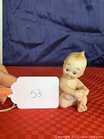 Front angled photo of a bisque porcelain Kewpie doll figurine seated, hands touching feet, with painted facial and hair details.