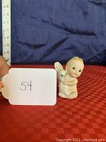 Kewpie doll and bunny figurine with measuring ruler showing size approximately 3 inches tall, placed on red textured cloth with blue background