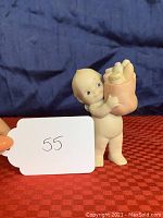 Front and side angle of bisque porcelain Kewpie doll holding brown paper grocery bag containing groceries including a corn carton.