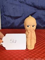 Front view of light peach colored Kewpie doll made of vinyl or rubber, seated with hands under chin and smiling face with detailed eyes