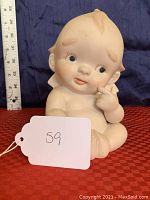 Front and partial side view of bisque porcelain Kewpie doll figurine, showing detailed facial features, hand-painted eyes, and seated pose.