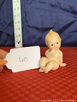 Front and side views of a bisque porcelain Kewpie doll figurine sitting on a red textured surface, next to a ruler for scale showing about 3 inches tall.