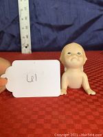 Front view of small bisque porcelain Kewpie doll figurine on red checkered fabric with measurement ruler showing approximately 3 inches tall.