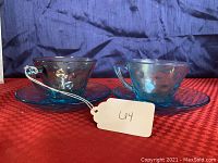 Pair of blue glass teacups with matching saucers on red surface with blue background. Light blue with raised polka dots pattern and clear handles visible.