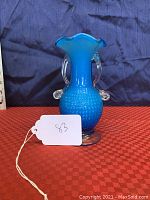 Front view of the blue blown cased glass flower vase with clear handles and hobnail texture on the body on a red cloth.