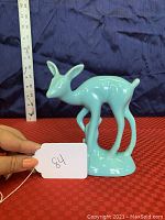 Side view of the green ceramic deer/fawn statue, showing height against a ruler, approximately 7.5 inches tall, on red textured fabric background.