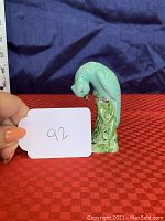 Front view of turquoise parrot figurine with green leafy base on a red checkered cloth, with a measuring tape in background for scale and a card labeled '92'.
