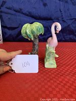 Front view of vintage flamingo and palm tree salt and pepper shakers on red surface with blue background, showing detailed hand-painted ceramic finish.