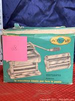 Photo of original green box showing front and side views of Atlas Model 150 Lusso pasta maker with hand crank and cutter attachment, showing some wear on the box.