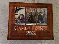 Front view of original box for 2017 Game of Thrones tarot card deck and guidebook showing three sample tarot cards ('The Hermit', 'Queen of Swords', 'The Empress') artwork on the lid.