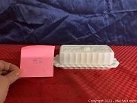 Side view of white milk glass butter dish with lid on red and blue fabric background, showing the scalloped base and paneled grape pattern.