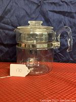 Front view of clear glass Pyrex Flameware 9 cup coffee percolator with lid, curved handle, and metal band on upper section, placed on red and black surface.