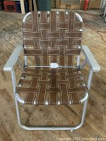 Front view of vintage aluminum folding lawn chair with brown woven webbing seat and backrest on concrete floor.