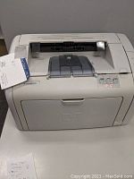 Front view of an off-white HP Laser Printer Model 1018 with paper output tray and a tag attached on the left side.