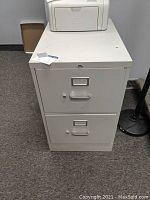 Front view of the grey two drawer metal filing cabinet showing two drawers with metal handles and label holders, with a white printer placed on top.