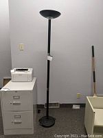 Full view of the black metal floor lamp standing next to a filing cabinet and broom, showing its height and overall design.