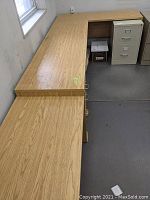 Photo showing left and center sections of brown woodgrain laminate corner desk, also partially showing metal file cabinet.
