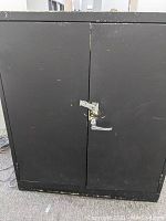 Closed black metal cabinet showing front doors with a handle and lock area for padlock.