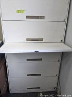 Close up view of part-open drawer in light grey metal lateral file cabinet, showing drawer face and recessed handle with label holder, locked and no key available
