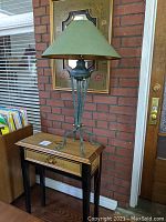 View of small decorative wooden table with floral painted top and drawer, black legs, with greenish metal lamp on top against brick wall