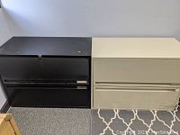 Two lateral file cabinets side by side, one black and one cream, showing exterior front and top surfaces and wear.