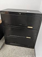 Front view of the closed black metal lateral file cabinet showing three drop-down drawer fronts with handles and lock cylinders.